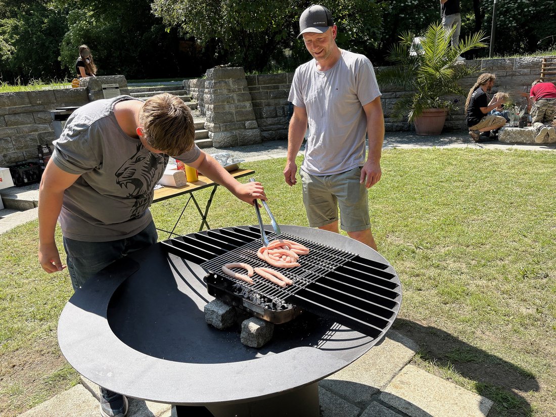Auf dem Bild ist ein großer Grill mit Würsten im Grünen zu sehen, zwei Personen stehen daneben.