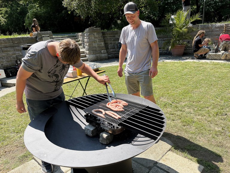 Auf dem Bild ist ein großer Grill mit Würsten im Grünen zu sehen, zwei Personen stehen daneben.