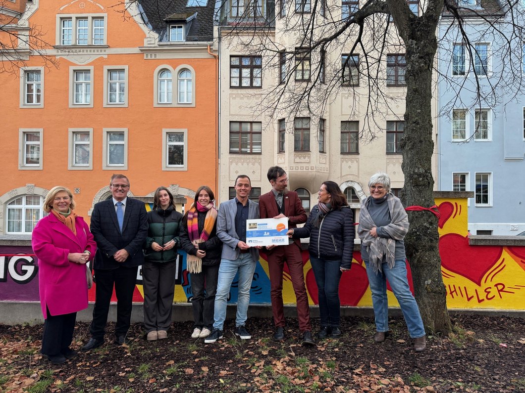 Auf dem Bild sind acht Personen vor der bunten Schulmauer zu sehen, zwei halten ein Schild der VR Bank mit einem Geldbetrag hoch.