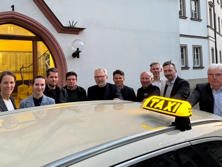 Auf dem Bild ist das Dach eines Taxis zu sehen, das vor dem Rathaus steht. Dahinter stehen nebeneinander 10 Personen.