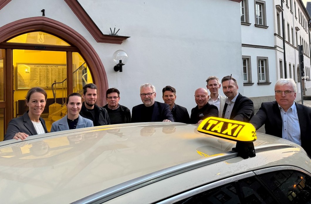 Auf dem Bild ist das Dach eines Taxis zu sehen, das vor dem Rathaus steht. Dahinter stehen nebeneinander 10 Personen.