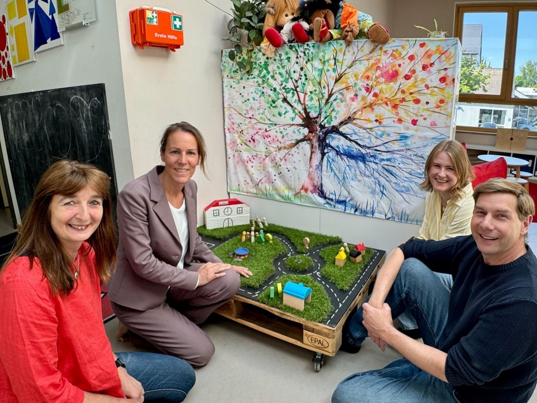 Auf dem Bild sind vier Personen zu sehen, die zwischen Spielsachen in einem Kindergarten am Boden sitzen.
