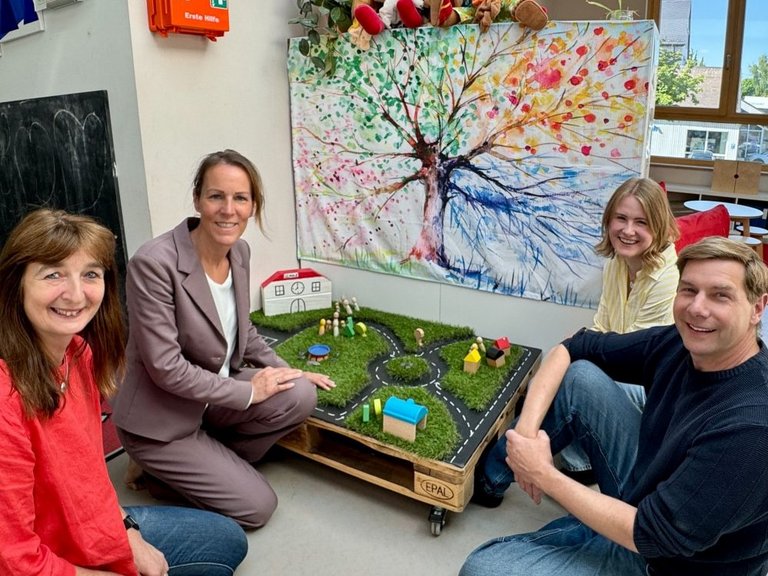 Auf dem Bild sind vier Personen zu sehen, die zwischen Spielsachen in einem Kindergarten am Boden sitzen.