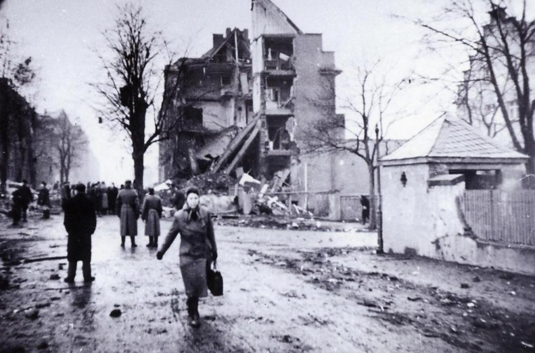 Auf dem Schwarz-Weiß-Bild sind Menschen zu sehen, die auf einer Straße laufen. Im Hintergrund völlig zerstörte Gebäude.