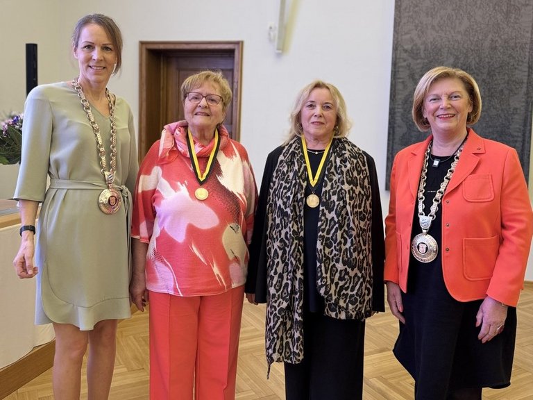 Auf dem Bild sind vier Frauen im Großen Sitzungssaal zu sehen. Die beiden äußeren tragen Amtsketten, die in der Mitte Ehrenmedaillen.
