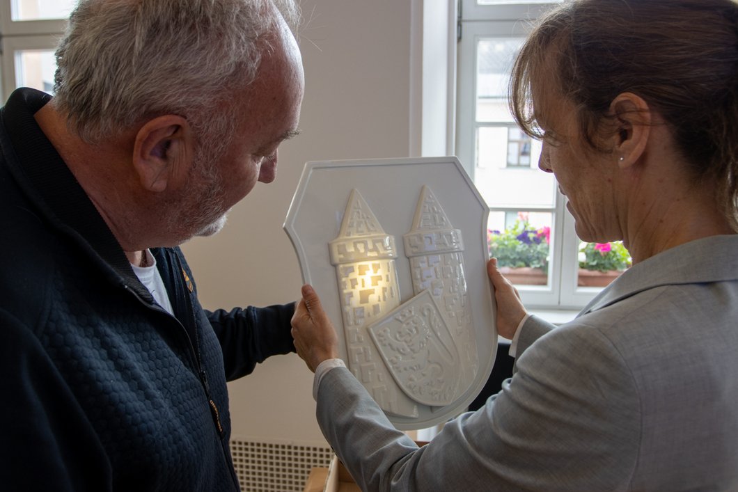 Auf dem Bild sind ein Mann und eine Frau zu sehen, die zusammen ein weißes Porzellan-Wappen halten, das von hinten beleuchtet ist.