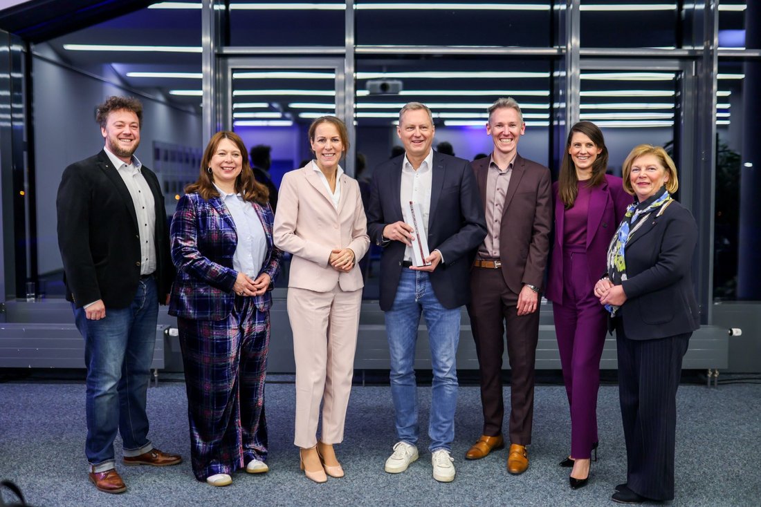Auf dem Bild sind sieben Personen in Businesskleidung zu sehen, die vor einer Glasfront stehen. Der Mann in der Mitte hält einen Preis in Händen.
