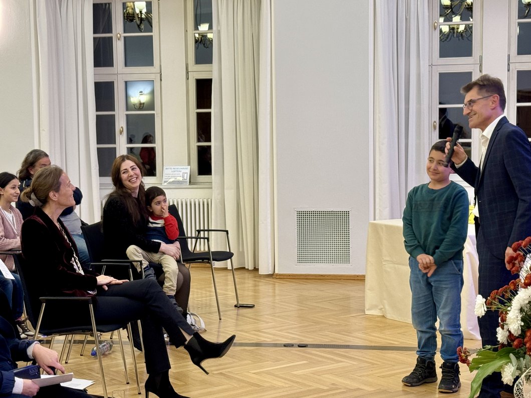 Auf dem Bild ist ein Mann mit Mikrofon zu sehen, der im Großen Saal spricht. Neben und vor ihm sind Kinder und zwei Frauen.