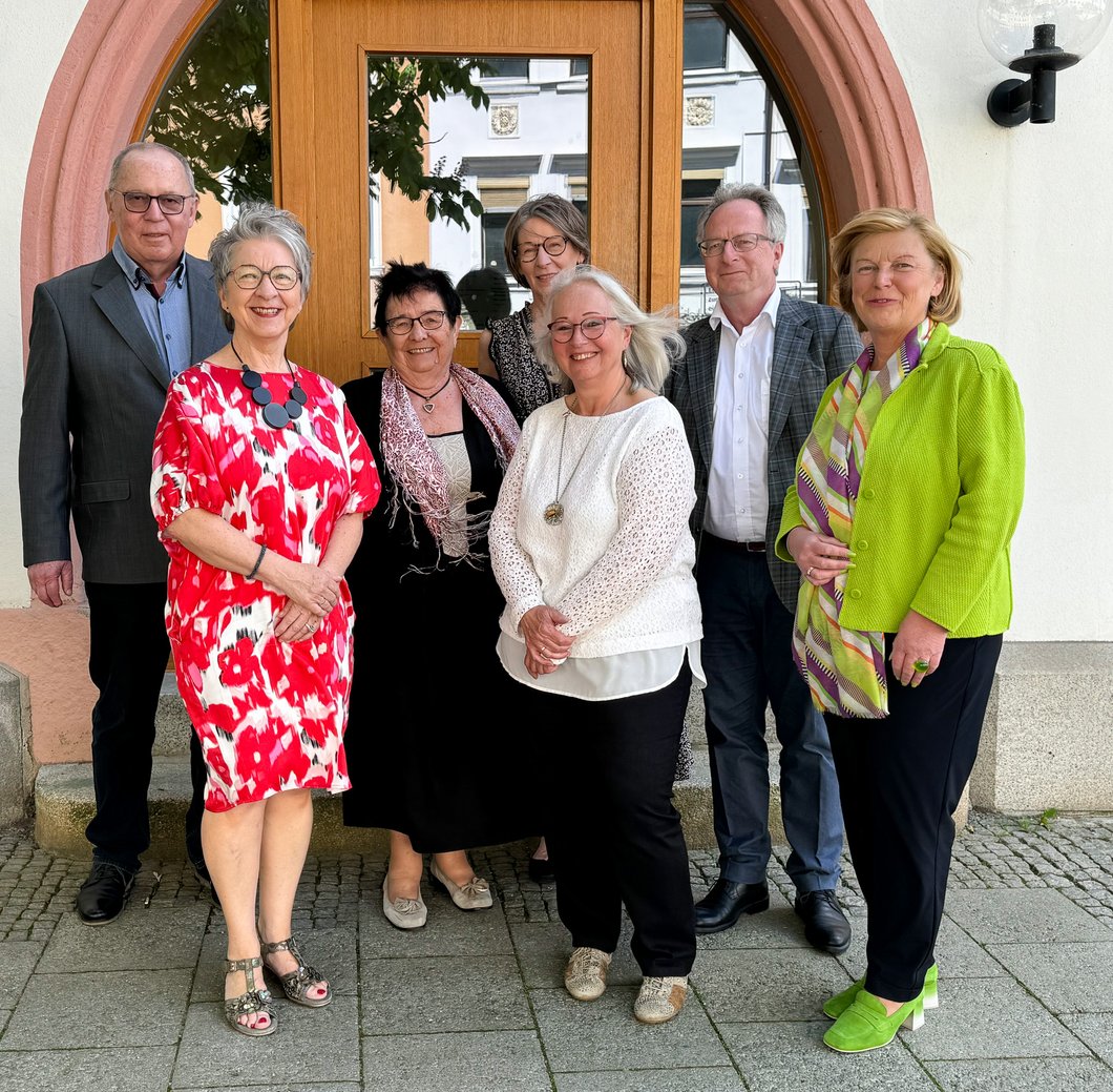 Auf dem Bild sind zwei Männer und fünf Frauen zu sehen, die draußen vor einer Tür stehen.