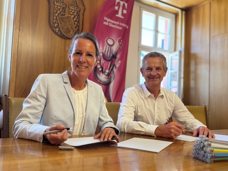 Auf dem Bild sind eine Frau und ein Mann in einem Saal zu sehen. Sie unterschreiben Dokumente.