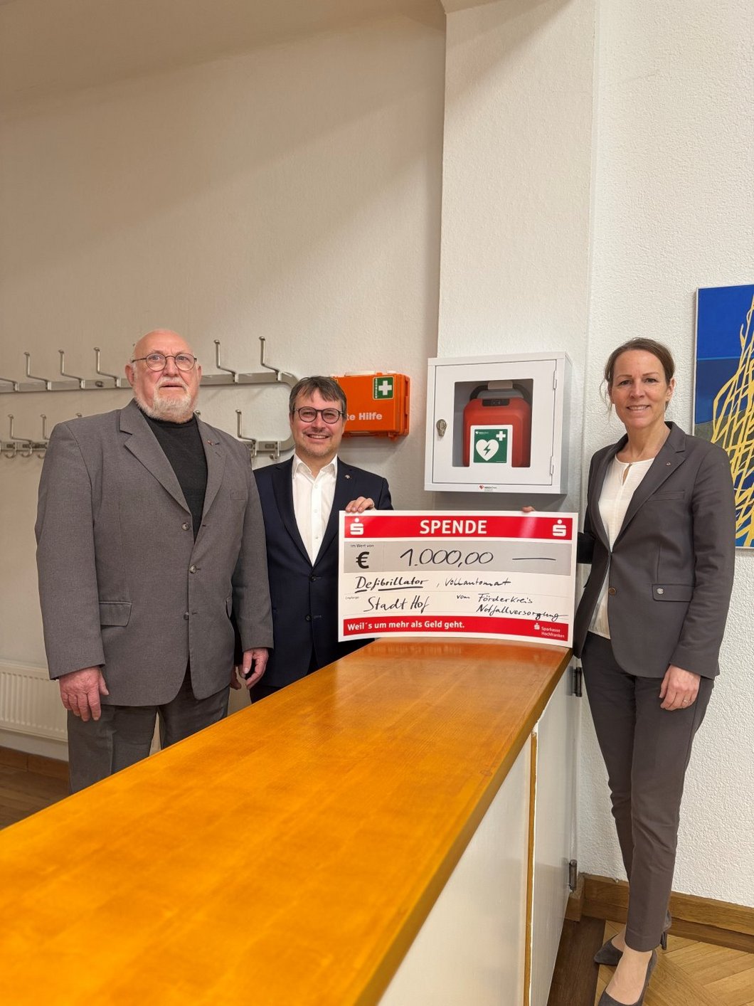 Auf dem Foto sind zwei Männer und eine Frau zu sehen, die mit einem großen Spendenscheck vor einem Defibrillator stehen.
