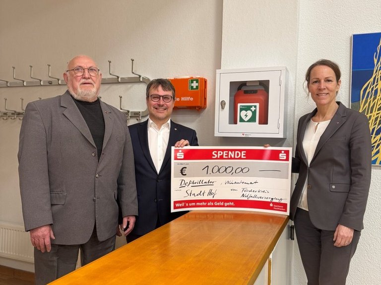 Auf dem Foto sind zwei Männer und eine Frau zu sehen, die mit einem großen Spendenscheck vor einem Defibrillator stehen.