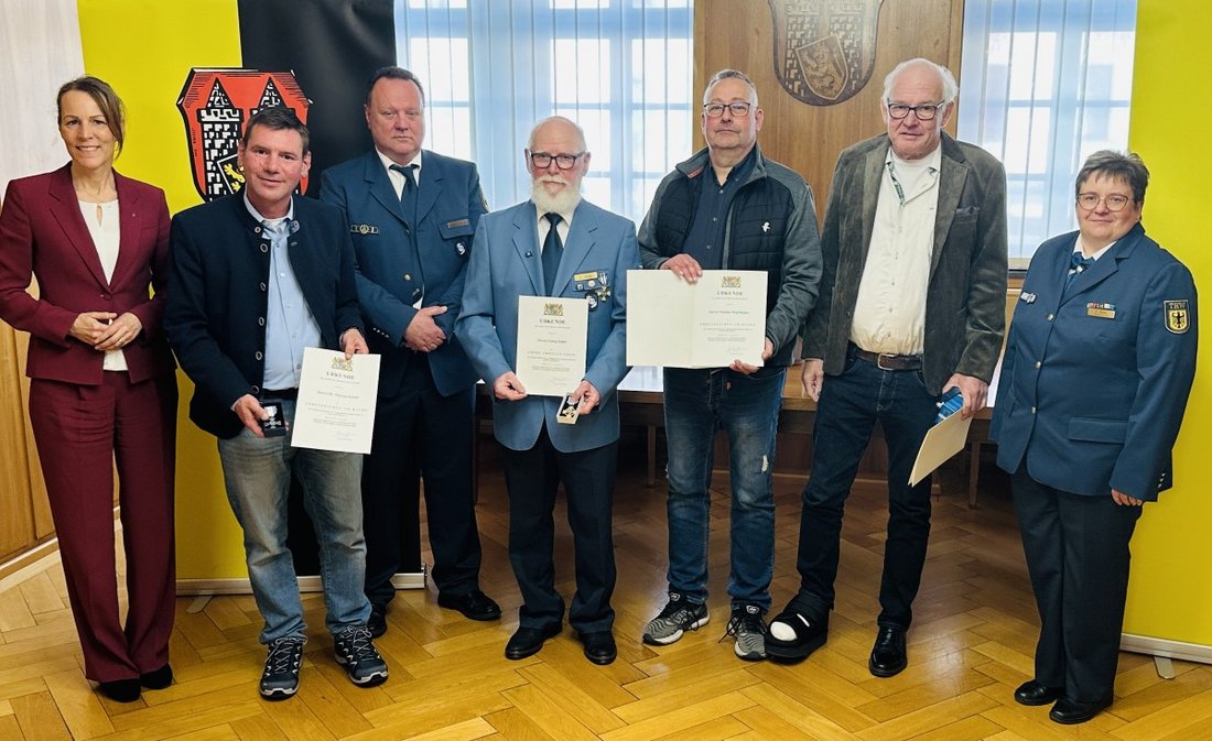Auf dem Bild sind sieben Personen in einem Saal vor dem Hofer Stadtwappen zu sehen. Vier von ihnen halten Urkunden.