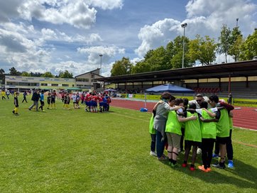 Auf dem Bild sind Kinder-Fußballteams in einem Stadion zu sehen, die sich im Kreis mit ihrem Trainer besprechen.
