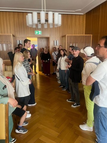 Über 20 Personen stehen in zwei Reihen in einem Saal und hören einem Mann zu.