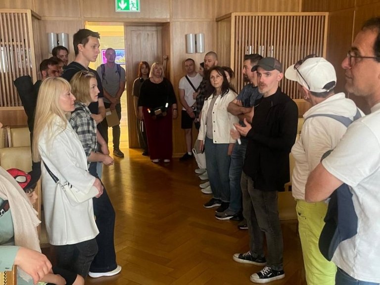 Über 20 Personen stehen in zwei Reihen in einem Saal und hören einem Mann zu.