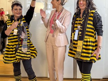 Auf dem Bild ist die Oberbürgermeisterin mit zwei als Bienen verkleideten Frauen zu sehen. Eine hält ein Stück abgeschnittene Krawatte hoch
