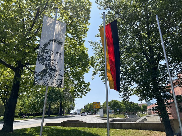 Auf dem Bild sind eine Deutschlandflagge und eine Flagge mit einem Gesicht zu sehen. Dahinter ein flaches Denkmal.