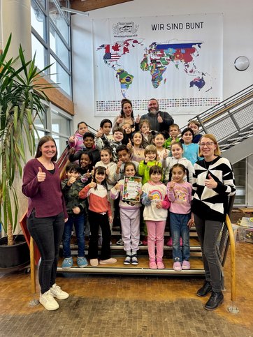 Auf dem Gruppenbild steht eine Schulklasse auf einer Treppe und zeigt Daumen hoch. Ein kleines Mädchen hält einen Gutschein. Dabei sind vier Erwachsene.