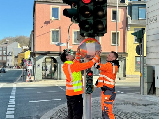 Auf dem Bild sind zwei Männer in oranger Kleidung zu sehen, die einen Spiegel an einer Ampel anbringen.