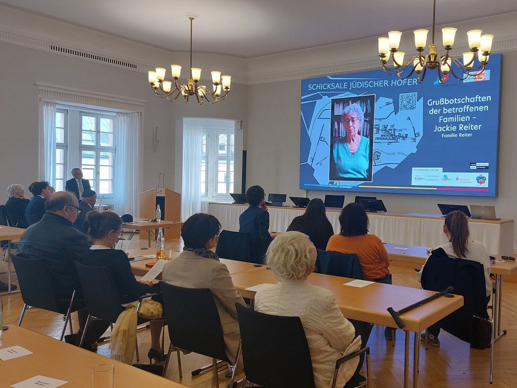 Auf dem Bild sind Menschen in einem Saal zu sehen, die eine Präsentation auf einem Monitor ansehen. Auf dem Monitor ist eine alte Frau zu sehen.