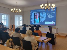 Auf dem Bild sind Menschen in einem Saal zu sehen, die eine Präsentation auf einem Monitor ansehen. Auf dem Monitor ist eine alte Frau zu sehen.