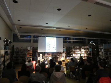 Auf dem Bild ist ein Publikum in einer Bibliothek zu sehen, das einem Mann mit Ukulele lauscht.