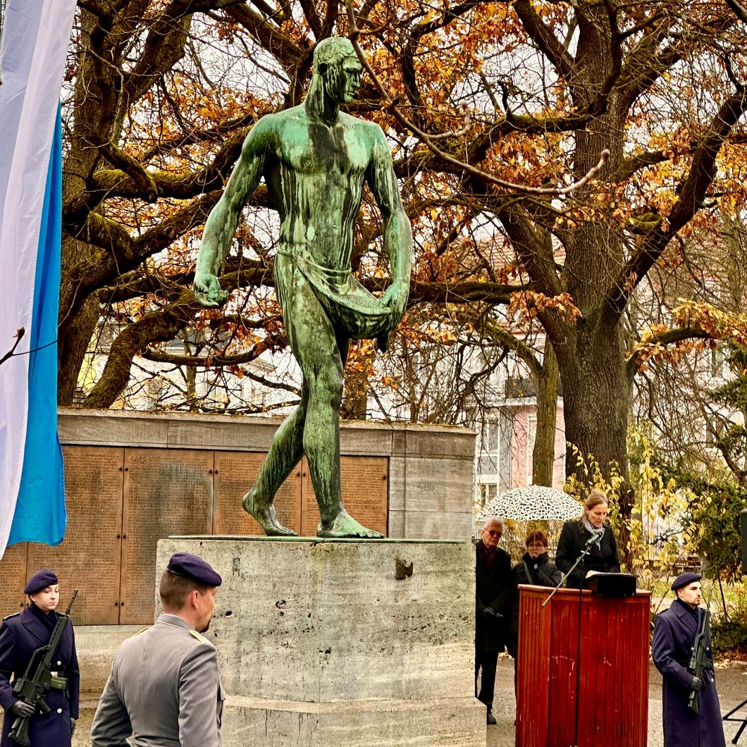 Auf dem Bild ist das Denkmal für die Gefallenen des 1. Weltkriegs im Wittelsbacherpark zu sehen. Am Pult davor hält Eva Döhla eine Rede, umgeben von Bundeswehrsoldaten.