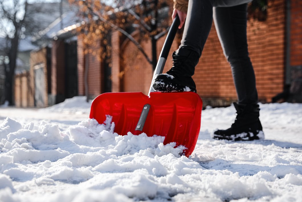 Auf dem Bild ist eine Person zu sehen, die mit einer roten Schaufel vor einem Gebäude Schnee schippt.