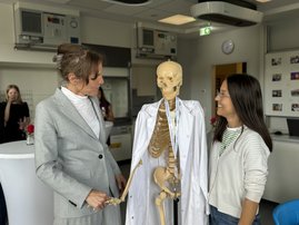 Auf dem Bild sind zwei Frauen zu sehen, zwischen denen ein Skelett steht, um das ein weißer Kittel hängt.