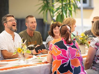Auf dem Foto sind sieben Personen zu sehen, die an einer gedeckten Tafel sitzen. Ein Mann lacht.