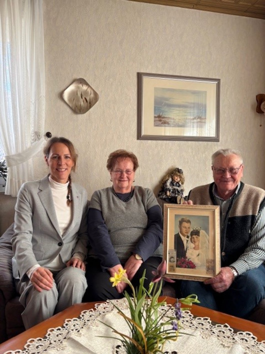 Auf dem Bild ist einälteres Ehepaar mit der Oberbürgermeisterin zu sehen. Sie sitzen in einem Wohnzimmer, der Ehemann hält ein altes gerahmtes Hochzeitsfoto.