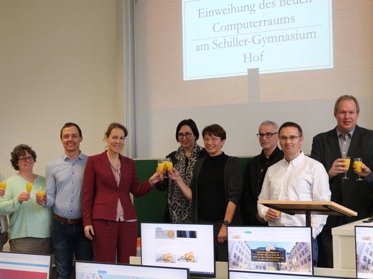 Auf dem Bild sind neun Personen zu sehen, die hinter einer Reihe Computer stehen und anstoßen.