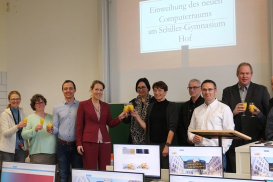 Auf dem Bild sind neun Personen zu sehen, die hinter einer Reihe Computer stehen und anstoßen.