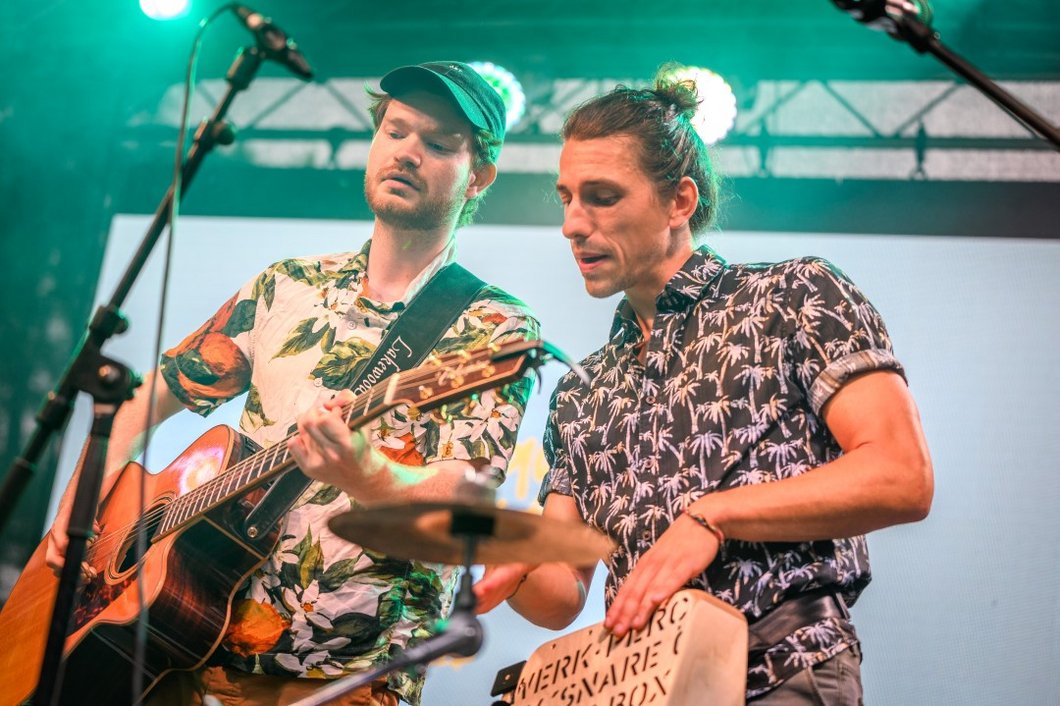 Auf dem Foto sind zwei Männer mit Musikinstrumenten zu sehen, die auf einer Bühne stehen.
