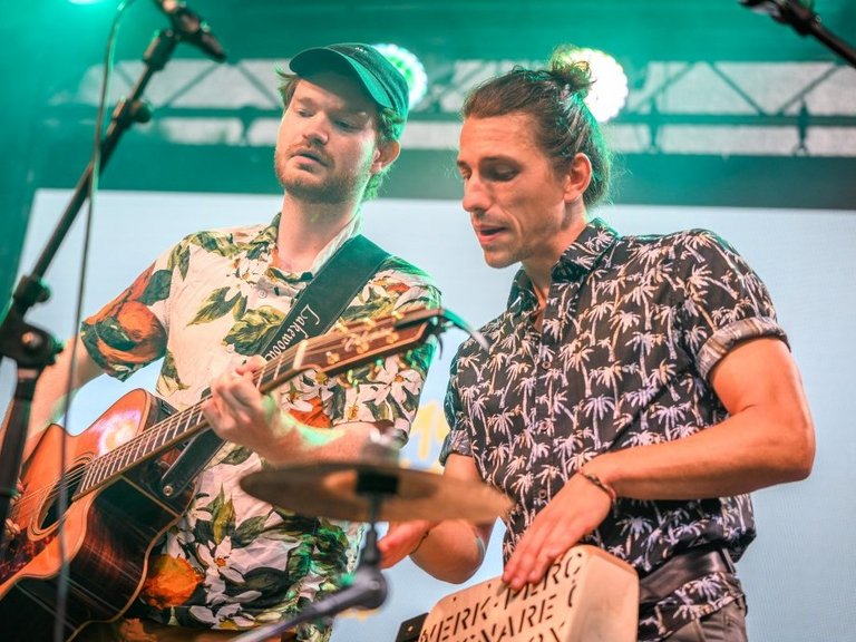 Auf dem Foto sind zwei Männer mit Musikinstrumenten zu sehen, die auf einer Bühne stehen.