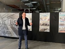 Auf dem Bild ist ein gestikulierender Mann zu sehen, der vor einer Stellwand mit farbigen Bildern steht. Der Innenraum dahinter ist eine Baustelle.