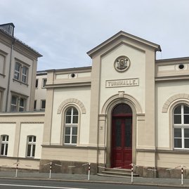 Auf dem Bild ist ein sandfarbenes Gebäude zu sehen mit der Aufschrift "Turnhalle""