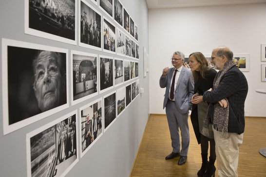 Auf dem Bild sind drei Personen zu sehen, die Schwarz-Weiß-Fotografien an einer Wand betrachten.
