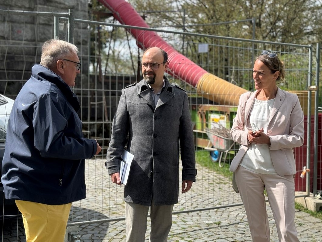 Auf dem Bild sind drei Personen zu sehen, die vor einer abgesperrten Baustelle stehen.