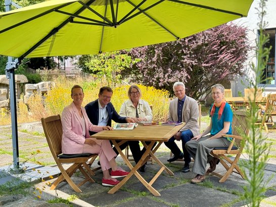 Auf dem Bild sind fünf Personen zu sehen, die unter einem Sonnenschirm an einem Holztisch sitzen.