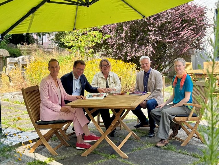 Auf dem Bild sind fünf Personen zu sehen, die unter einem Sonnenschirm an einem Holztisch sitzen.