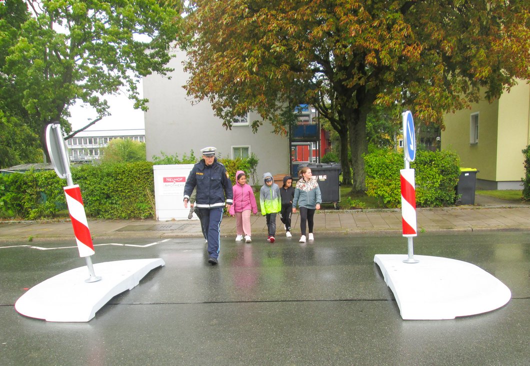 Auf dem Bild ist eine Polizistin zu sehen, die an der Querungshilfe mit vier Kindern über die Straße geht.