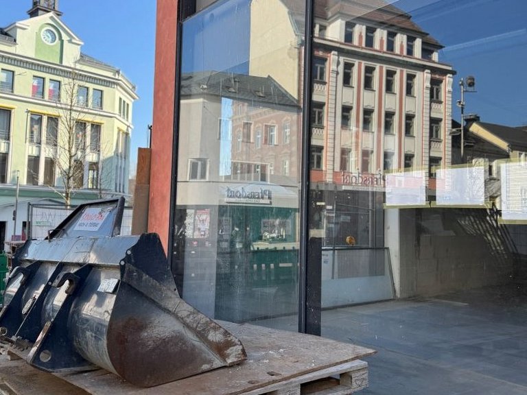 Auf dem Bild ist das Schaufenster eines Gebäudes von außen zu sehen, davor Baugeräte und Paletten.