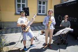 Auf dem Bild sind drei Musiker zu sehen, die live Gitarre, Saxofon und Keyboard auf einem Marktplatz spielen.