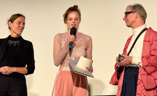 Auf dem Bild sind zwei Frauen und ein Mann zu sehen. Eine der Frauen hält einen Preis in der Hand und spricht in ein Mikrofon.