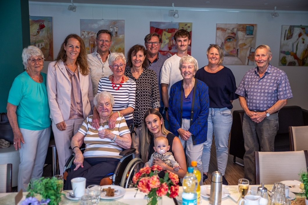 Auf dem Bild ist eine Gruppe von jungen und alten Menschen hinter einer geschmückten Tafel zu sehen. Eine alte Dame im Rollstuhl sitzt vorne.