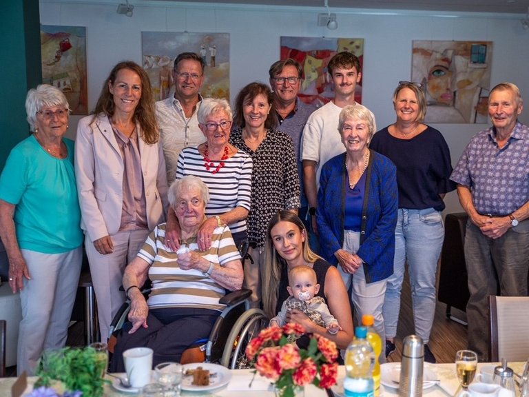 Auf dem Bild ist eine Gruppe von jungen und alten Menschen hinter einer geschmückten Tafel zu sehen. Eine alte Dame im Rollstuhl sitzt vorne.