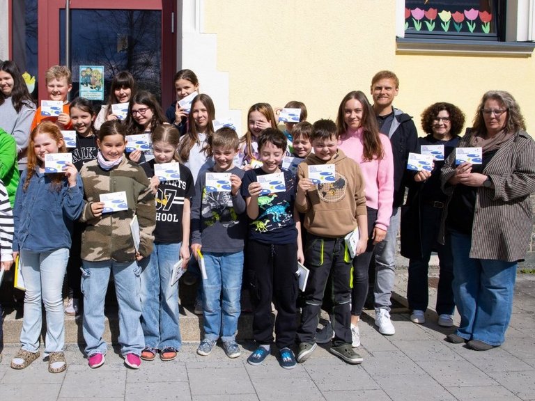 Auf dem Foto ist eine Schulklasse zu sehen, die vor der Schule steht. Die Kinder halten das Osterferienprogramm hoch.