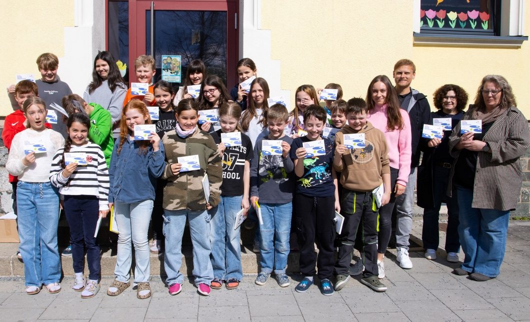 Auf dem Foto ist eine Schulklasse zu sehen, die vor der Schule steht. Die Kinder halten das Osterferienprogramm hoch.